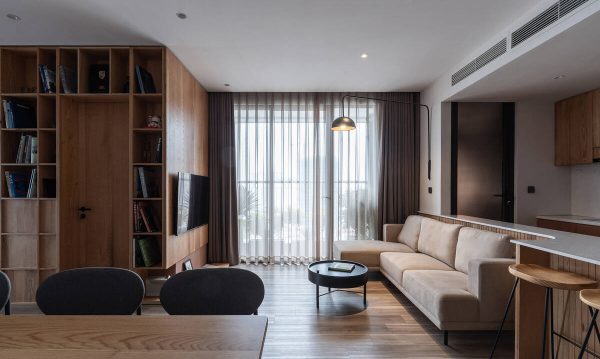 The living room area with the kitchen partition is removed to bring a more spacious and airy space. In this view, you can see a lot of indoor furniture made of wood such as wooden floors, wooden dining tables, wooden chairs, wooden bookshelves, and wooden kitchen cabinets.